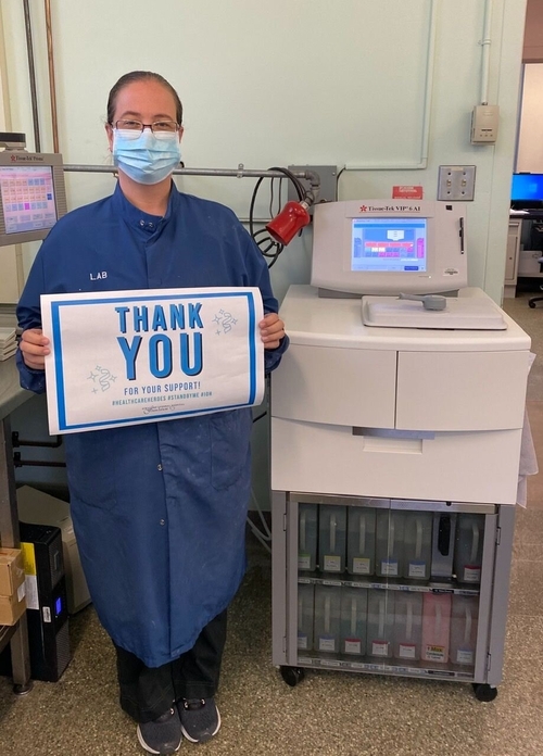 A lab technician wearing a blue lab coat, face mask, and glasses stands beside a tissue processor in a hospital laboratory. She holds a sign that reads “Thank You for Your Support!” with hashtags #HealthcareHeroes and #StratfordProud. Monitors and lab equipment are visible in the background.